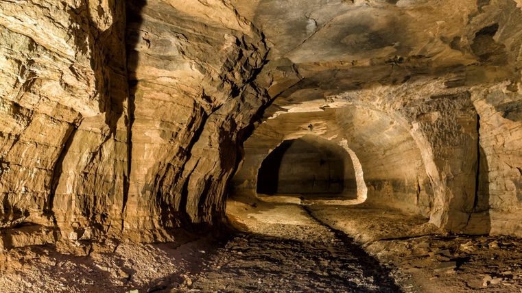 A illuminated tunnel with rocky walls and a vaulted ceiling. The floor is uneven and covered with debris. In the background, an opening is visible.