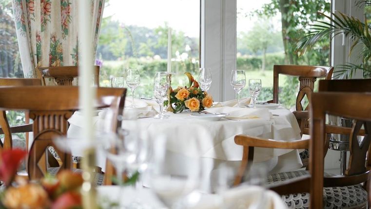 Ein eleganter Tisch in einem hellen Raum mit Blick ins Grüne. Der Tisch ist mit einer blütenförmigen Dekoration und Gläsern gedeckt.