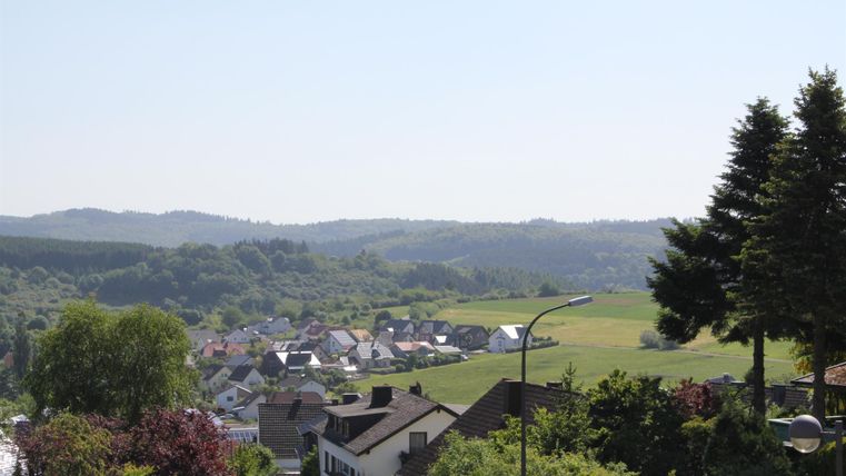 Eine ruhige Landschaft mit einem kleinen Dorf und sanften Hügeln im Hintergrund. Der Himmel ist klar und die Umgebung ist grün und lebendig.
