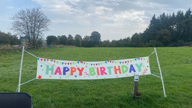 Ein bunter Geburtstagsbanner steht in einer Wiese. Im Hintergrund sind Bäume und ein bewölkter Himmel zu sehen.
