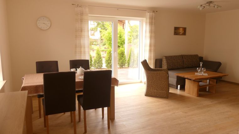 Ein helles Wohnzimmer mit einem Esstisch und Stühlen. Auf der gegenüberliegenden Seite befindet sich eine gemütliche Sitzecke und große Fenster mit Blick ins Freie.