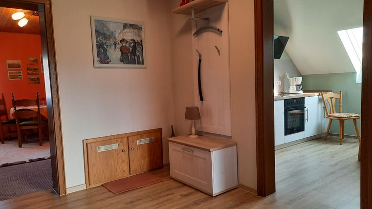 A bright hallway with wooden flooring and a small seating area. In the background, you can see an open kitchen corner with a cooking area.