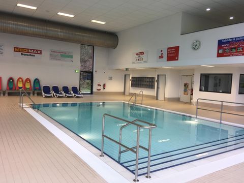 A bright indoor swimming pool with clear water and a ladder. There are loungers by the edge of the pool.