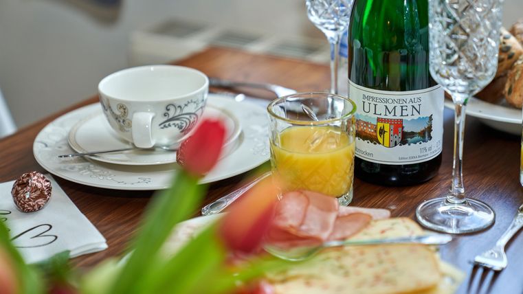 A lovingly set table with a cup, orange juice, sparkling wine, and fresh bread. In the foreground, tulips can be seen.