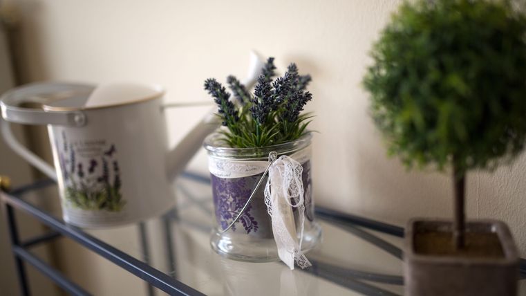 Ein dekorativer Pflanzenbehälter mit Lavendel und einer Schleife steht auf einem Tisch. Nebenan befindet sich ein silbernes Gießkanne und ein kleiner, runder Topf mit einer grünen Pflanzen.