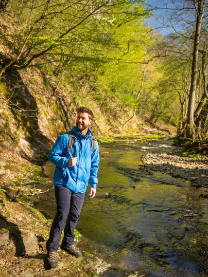 Ein Mann steht am Ufer eines klaren Baches in einem grünen Wald. Es ist ein sonniger Tag und die Umgebung wirkt einladend und freundlich.