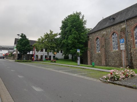 Een rustige straat met groene bomen en een oude kerk. Links zijn enkele gebouwen en bloemperken te zien.