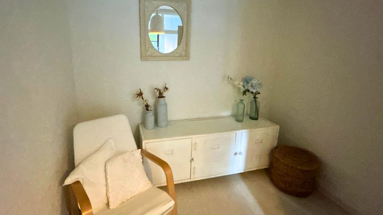 A bright room with a cozy chair and a small table. On the table, there are vases with flowers, and a mirror is hanging on the wall.