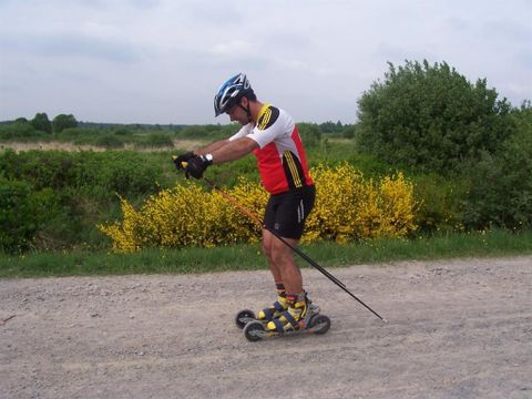 Ein Mann fährt auf einem Rollskiweg und trägt Sportkleidung sowie einen Helm. Im Hintergrund sind grüne Sträucher und gelbe Blumen zu sehen.