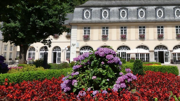Een mooi bloemenbed met rode en paarse bloemen voor een elegant gebouw. De omgeving is groen en uitnodigend.