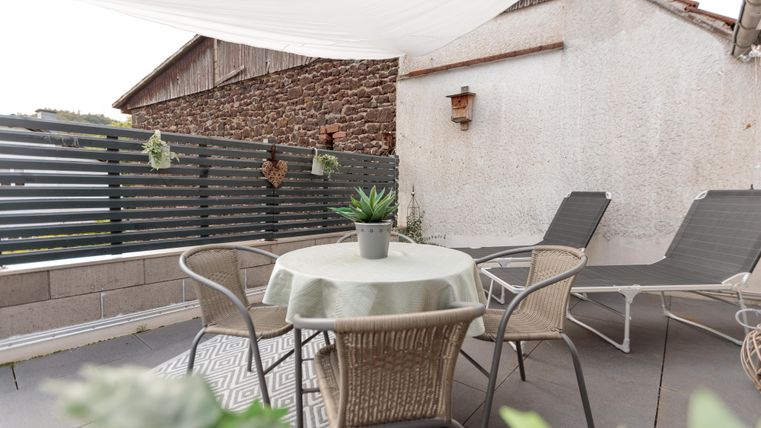 Eine gemütliche Terrasse mit einem Tisch und Stühlen. Im Hintergrund sind eine Wand und eine Sonnenschutzplane zu sehen.