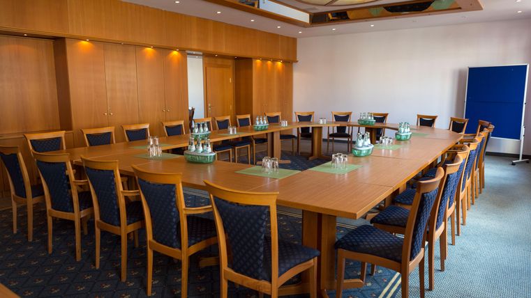 Ein moderner Konferenzraum mit einem großen Tisch und mehreren Stühlen. Auf dem Tisch stehen Wasserkaraffen und Gläser, und die Wände sind in neutralen Farben gehalten.