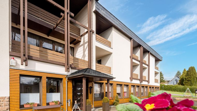A modern building with wooden cladding and several balconies. In the foreground, there are flowers and an inviting front door.