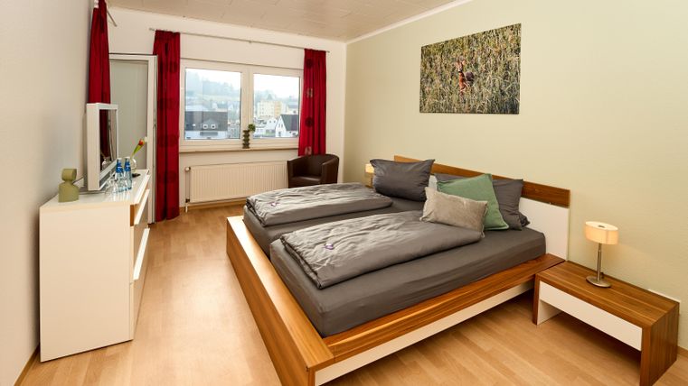 Ein gemütliches Schlafzimmer mit einem großen Bett und modernen Möbeln. Helle Wände und rote Vorhänge sorgen für eine freundliche Atmosphäre.
