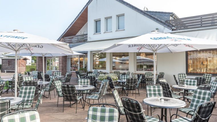 A terrace with many tables and chairs, surrounded by umbrellas. In the background, a modern building can be seen.