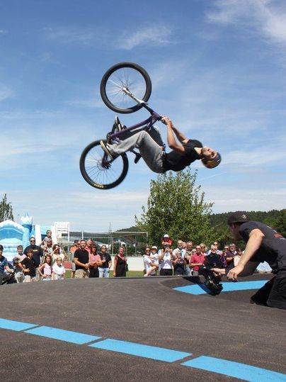 Ein BMX-Fahrer macht einen spektakulären Rückwärtssalto über eine Rampe. Im Hintergrund versammelt sich eine enthusiastische Menge, die die Aufführung verfolgt.