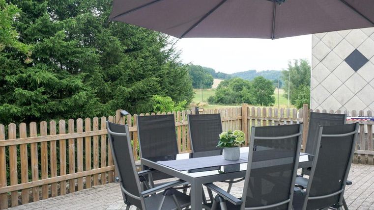 Ein einladender Terrassentisch mit sechs Stühlen und einem Sonnenschirm. Im Hintergrund sind Bäume und eine Wiese zu sehen.