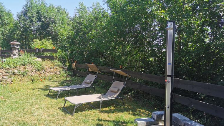 Een rustige tuinruimte met twee ligbedden en een buitendouche. Omgeven door bomen en een houten hek.