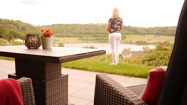 A woman stands on a terrace and looks out at a green landscape with a body of water in the background. The table is decorated with a flower vase and a decorative basket.