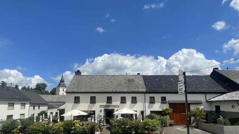 Außenansicht der Gastwirtschaft Sünnen mit Terrasse und Sonnenschirmen, umgeben von Pflanzen, unter blauem Himmel mit Wolken.