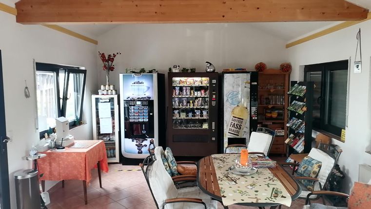 Ein gemütlicher Raum mit mehreren Stehtischen und Stühlen. An den Wänden stehen Verkaufsautomaten und ein Tisch ist mit einer Tischdecke dekoriert.