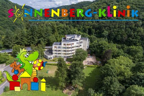 A modern building of the Sonnenberg Clinic surrounded by green nature. In the foreground, a colorful children's logo can be seen.