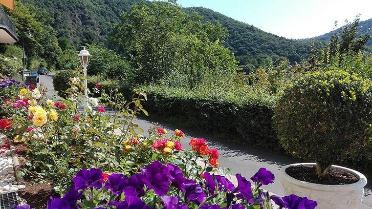 Eine blühende Gartenlandschaft mit bunten Blumen und einem gepflegten Weg. Im Hintergrund sind sanfte Hügel und Bäume zu sehen.