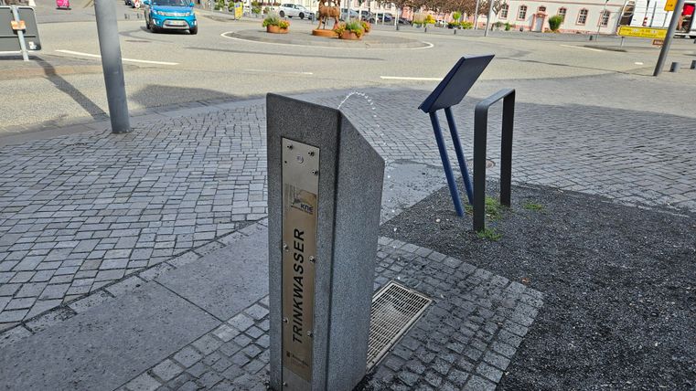 A drinking water fountain made of stone stands in a paved square. In the background, cars and a sign are visible.