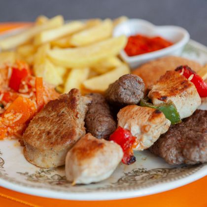 Ein Teller mit gegrilltem Fleisch, Pommes, Salat und roter Sauce. Verschiedene Fleischsorten und Gemüse sind zu sehen.