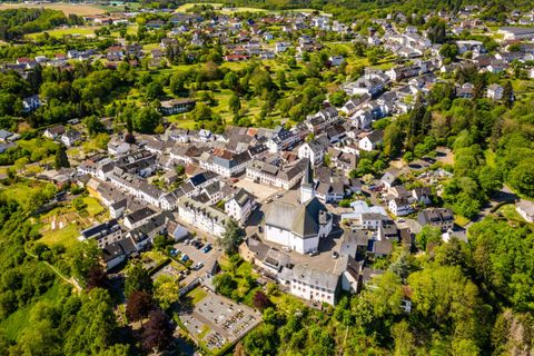 Eine malerische Ansicht eines kleinen Dorfes, umgeben von grünen Wäldern und Feldern. Die ordentlichen Straßen und Gebäude verleihen dem Ort einen charmanten Charakter.