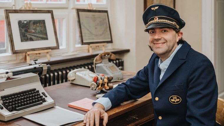 Ein Mann in einer blauen Uniform mit einer Mütze sitzt an einem Schreibtisch mit einer Schreibmaschine und zwei Telefonen.