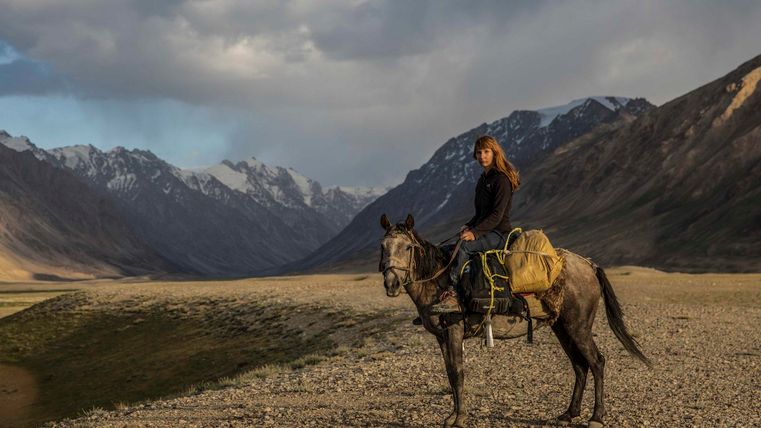 Een ruiter zit op een grijs paard in een bergachtig landschap. Majestueuze bergen en een dramatische lucht omringen haar.