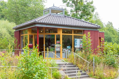 Ein modernes, rot gestrichenes Gebäude mit großen Fenstern und einem Glasdach. Umgeben von grünen Pflanzen und einem gepflegten Eingang.