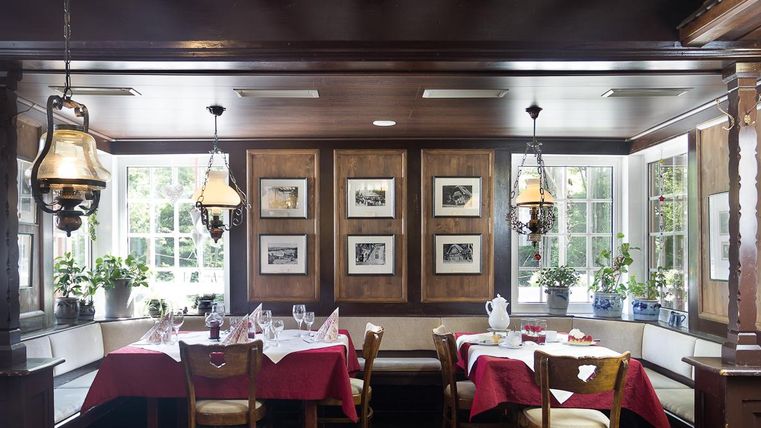 Ein gemütliches Restaurant mit Holzmöbeln und einer warmen Atmosphäre. Tische sind mit roten Tischdecken gedeckt und große Fenster bieten viel Tageslicht.