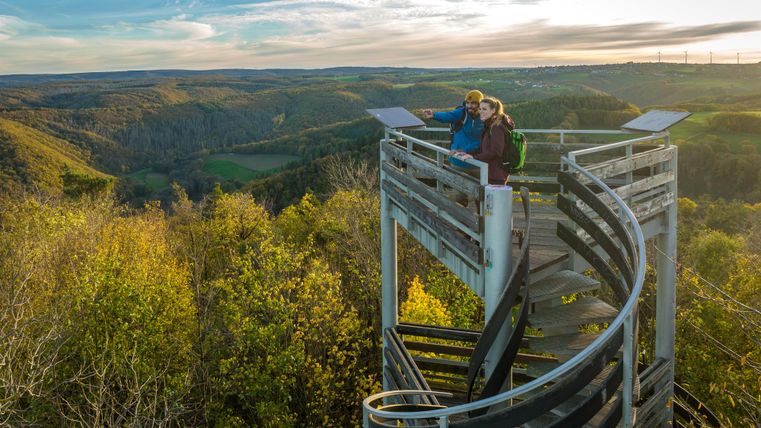 Zwei Personen stehen auf einer Aussichtsplattform und genießen die Landschaft. Im Hintergrund sind sanfte Hügel und ein schöner Himmel zu sehen.