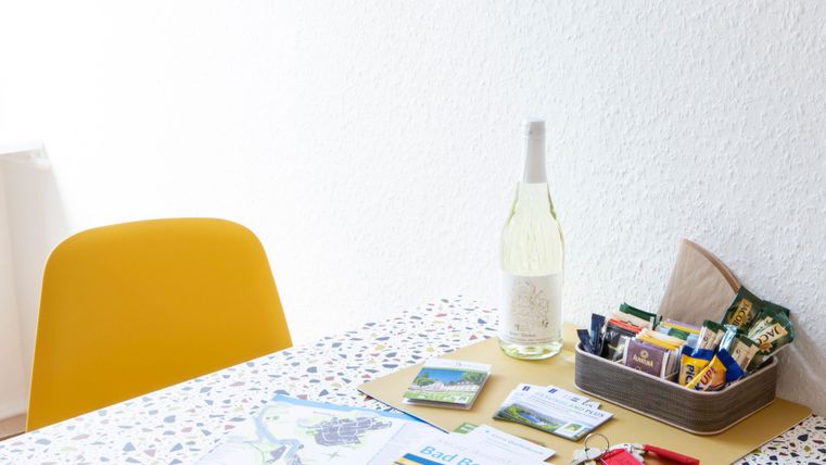 Ein einladender Tisch mit einem gelben Stuhl, einer Flasche Wein und verschiedenen Informationsmaterialien. Auf dem Tisch liegen auch Snacks und Schlüssel.