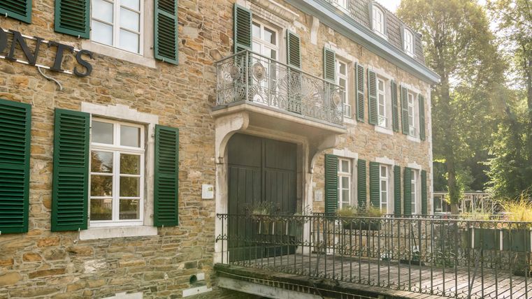 Een historisch stenen gebouw met groene luiken en een balkon. Een kleine houten brug leidt naar de ingang.