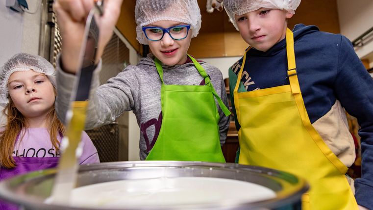 Drei Kinder in Kochschürzen und Haarnetzen stehen um einen Kochtopf. Sie rühren aufmerksam in die Mischung.