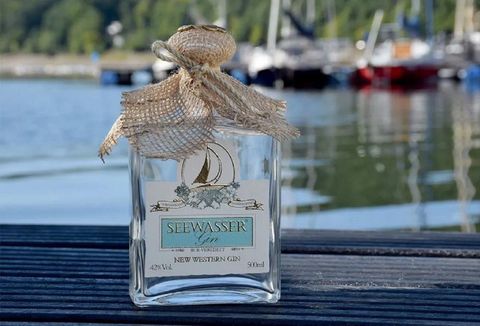 Eine Flasche Seewasser-Gin steht auf einem Holzsteg am Wasser. Im Hintergrund sind Boote und eine grüne Landschaft zu sehen.