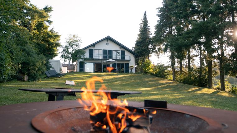 Ein schönes Haus umgeben von Bäumen und grünem Rasen. Im Vordergrund brennt ein Feuer in einem Grill.