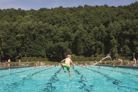 A swimming pool with a boy jumping into the water. In the background, other swimmers and green trees can be seen.