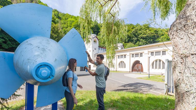 Een groot, blauw schaufelrad staat op de voorgrond, terwijl twee personen ervoor staan en met elkaar praten. Op de achtergrond zijn historische gebouwen en veel groen te zien.