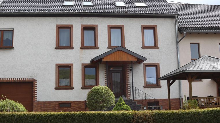 Ein modernes Wohnhaus mit einer hellen Fassade und braunen Fensterrahmen. Der Eingang ist mit einem überdachten Vorbau ausgestattet und von einem gepflegten Garten umgeben.