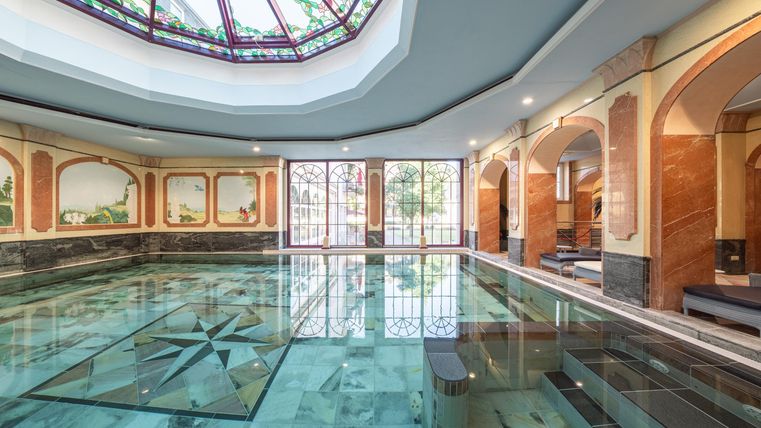 Een luxe zwembad met een elegant glazen dak. De muren zijn versierd met kunstzinnige schilderijen en de ruimte is licht en uitnodigend.