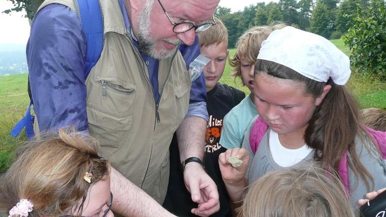 Ein Mann zeigt einer Gruppe von Kindern Steine. Sie stehen im Freien, umgeben von Bäumen und Wiesen.