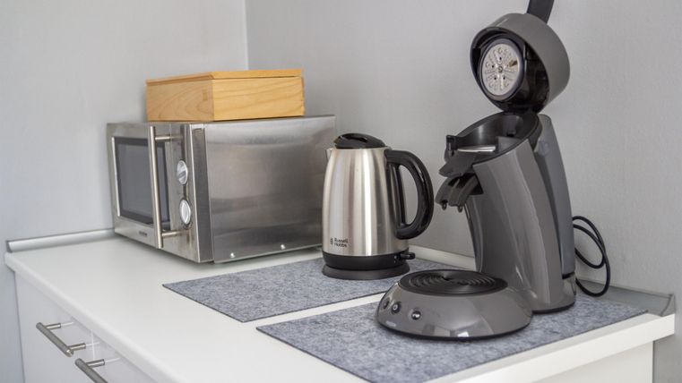 A modern kitchenette with a kettle, a coffee maker, and a microwave. The kitchen appliances are neatly arranged on a white table.
