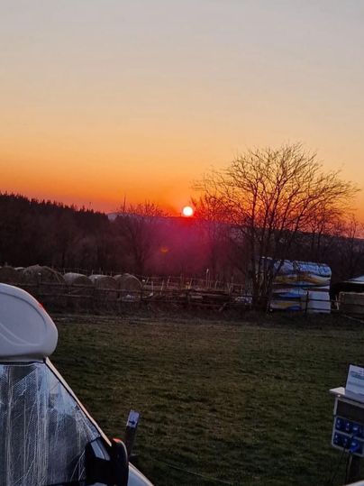 Ein schöner Sonnenuntergang über der Landschaft, mit leuchtenden Farben am Himmel. Im Vordergrund sind Bäume und eine Wiese zu sehen.