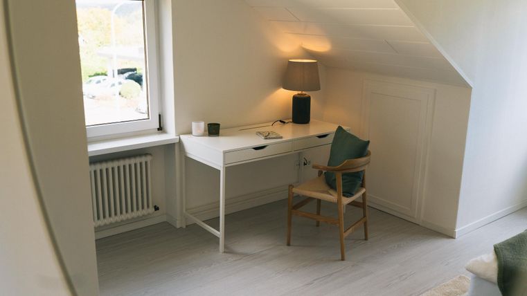 Een lichte werkplek met een witte bureau en een stoel staat onder een raam. Een lamp zorgt voor sfeervol licht in de ruimte.