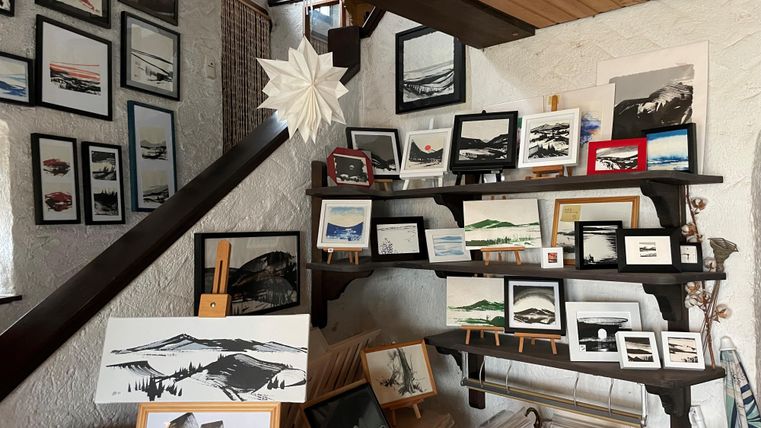 Eine Galerie mit verschiedenen Kunstwerken, darunter Bilder von Bergen und Landschaften. Die Werke hängen an der Wand und sind auf Regalen ausgestellt.