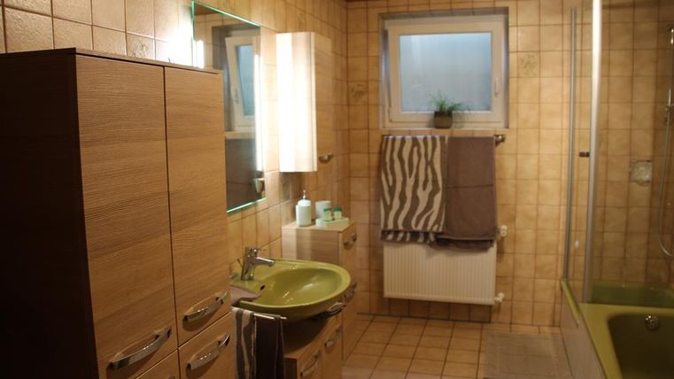 A modern bathroom with wooden furniture and a green sink. There is a window, a shower, and decorative towels.